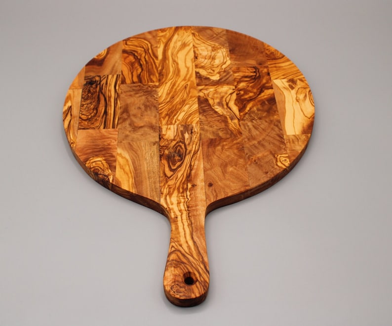 May include: A round olive wood cutting board with a handle. The board is made of multiple pieces of wood that have been joined together to create a unique pattern.