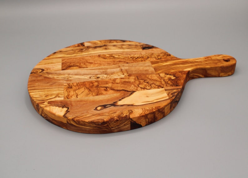 May include: A round olive wood cutting board with a handle. The board has a natural wood grain pattern and is perfect for serving cheese, bread, or other appetizers.
