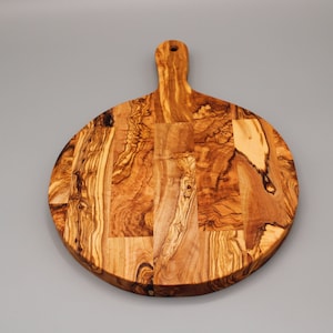 May include: Round olive wood cutting board with a handle. The board has a natural wood grain pattern and is made of multiple pieces of wood.
