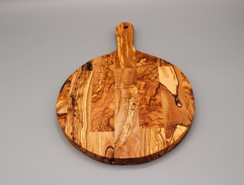 May include: Round olive wood cutting board with a handle. The board has a natural wood grain pattern and is made of multiple pieces of wood.