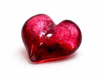 May include: A heart-shaped, ruby-red glass object. The translucent glass has a textured appearance, with darker red areas creating depth. The object is set against a white background, highlighting its shape and color. The heart is a symbol of love and affection.