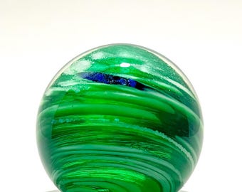 May include: A vibrant green glass sphere with swirling patterns of white and a touch of blue. The orb appears translucent, with light passing through the glass, creating a dynamic visual effect. The sphere sits on a white surface.