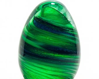 May include: A decorative glass paperweight in an egg shape. The paperweight features swirling patterns of green and dark blue, creating a dynamic and artistic design. The glass is smooth and glossy, reflecting light.
