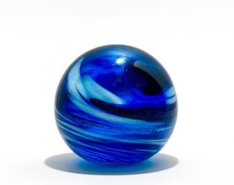 May include: A blue glass marble with swirling patterns of light blue and white. The sphere is highly reflective, with a glossy finish. The marble sits on a white surface, casting a soft shadow.