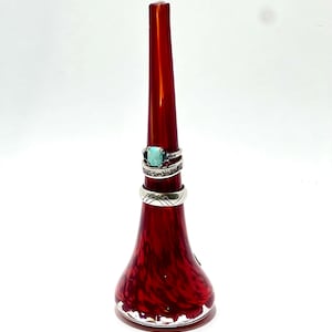 May include: A red glass ring holder with a flared base and a long, tapered top. Three rings with silver bands and gemstones are displayed on the holder. The holder has a speckled design.