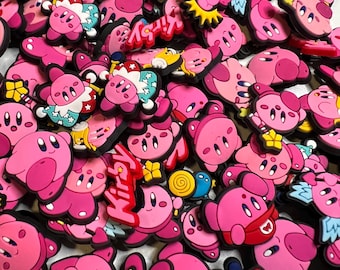 May include: A collection of pink Kirby character charms in various poses and designs. Some charms have additional colorful details, including a crown, a star, and the word "Kirby".