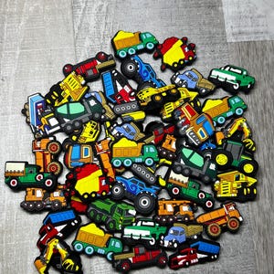 May include: A collection of colorful toy trucks and construction vehicles, including dump trucks, cement mixers, and excavators. The vehicles are in various colors like red, yellow, green, and blue, and are arranged in a pile.