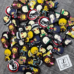 May include: A collection of colorful rubber charms featuring anime characters. The charms are in various shapes and sizes, with detailed facial features and clothing. Some charms have text and logos. A white label with the text "x tina creates" is also present.