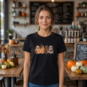 May include: A black t-shirt featuring a graphic print of four ghosts in a row. The ghosts have different patterns including orange pumpkins, a black witch hat, floral designs, and a purple plaid print, with a small bat on the far right ghost.