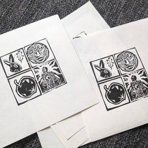 Può includere: Due stampe in linoleum in bianco e nero su carta bianca. Ogni stampa presenta quattro quadrati con immagini di un coniglio, un sole, una persona e un frutto.