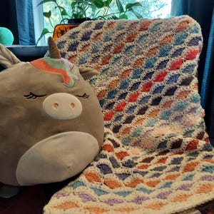May include: A colorful crocheted blanket with a diamond pattern in shades of blue, purple, orange, and pink. The blanket is draped over a chair with a plush unicorn toy sitting on it.