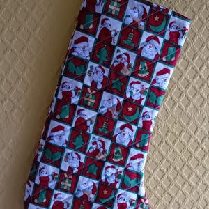 May include: Christmas stocking with a green hanging loop. The fabric features a pattern of red squares with images of Santa Claus, Christmas trees, and gifts on a white background. The stocking is quilted.