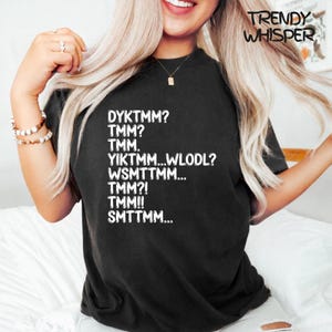 Può includere: T-shirt nera con testo bianco. Il testo include una serie di acronimi e il marchio "TRENDY WHISPER" in alto a destra. La maglietta ha un girocollo e maniche corte.