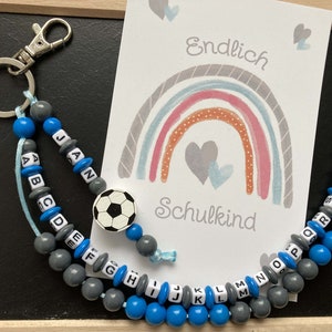 Könnte beinhalten: Eine blaue und graue Perlenkette mit einem Fußball-Anhänger. Die Perlen sind mit dem Alphabet und den Wörtern "Jan" und "Schulkind" beschriftet.