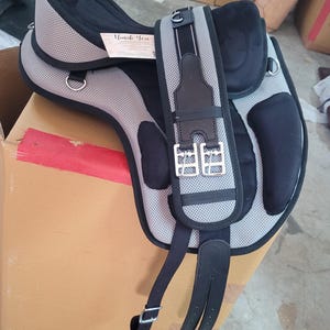 May include: A horse saddle featuring a grey mesh textured surface with black padded sections, silver metal D-rings, and a black girth strap with two silver buckles. A small rectangular tag with the text Thank You is attached to the saddle.