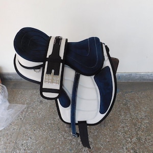 Puede incluir: Una silla de montar inglesa blanca y azul marino. La silla tiene un asiento y paneles acolchados, con herrajes negros y plateados. La silla está diseñada para la comodidad y el rendimiento.