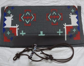 May include: A gray saddle pad with a geometric pattern in red, white, blue, and green. The pad has a brown leather flap and a brown leather strap with a braided rope detail.