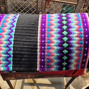 May include: A black and white saddle pad with a colorful geometric pattern in shades of pink, purple, turquoise, and white. The pad is woven with a textured design.