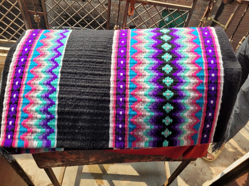 May include: A black and white saddle pad with a colorful geometric pattern in shades of pink, purple, turquoise, and white. The pad is woven with a textured design.