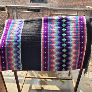 May include: A black and white saddle blanket with colorful geometric patterns in pink, purple, blue, and white. The blanket is woven with a fringe on the bottom edge.
