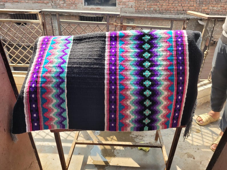 May include: A black and white saddle blanket with colorful geometric patterns in pink, purple, blue, and white. The blanket is woven with a fringe on the bottom edge.