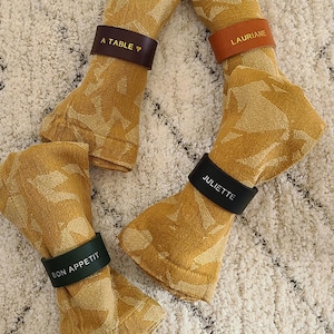 May include: Four folded mustard-yellow napkins with leaf patterns, each held by a leather napkin ring. The rings are brown, orange, black, and green, with text that reads "A TABLE", "LAURIANE", "JULIETTE", and "BON APPETIT".