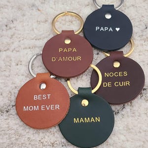 May include: A collection of six circular leather keychains in various colors including tan, brown, dark green, and navy blue. Each keychain features a gold or silver metal rivet and a metal ring, with text embossed in gold or white, reading PAPA, PAPA D'AMOUR, NOCES DE CUIR, BEST MOM EVER, and MAMAN.