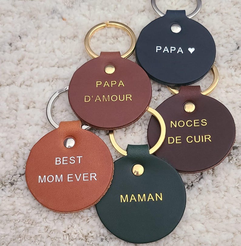 May include: A collection of six circular leather keychains in various colors including tan, brown, dark green, and navy blue. Each keychain features a gold or silver metal rivet and a metal ring, with text embossed in gold or white, reading PAPA, PAPA D'AMOUR, NOCES DE CUIR, BEST MOM EVER, and MAMAN.