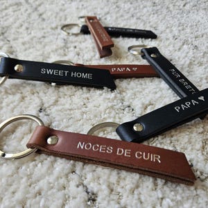 May include: A collection of leather keychains in various colors, including black and brown. Each keychain features a silver keyring and embossed text, such as "SWEET HOME" and "PAPA". The keychains are arranged on a textured, off-white surface.