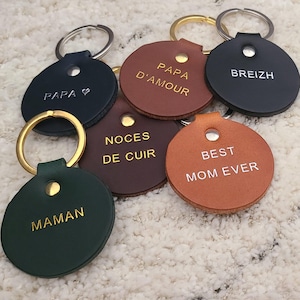 Puede incluir: Una colección de llaveros de cuero en varios colores, incluyendo verde, marrón y negro. Cada llavero tiene un diseño circular con un anillo metálico y está grabado con textos como "PAPA", "MAMAN" y "BEST MOM EVER".