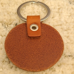 May include: A circular brown leather keychain featuring a textured surface and a silver metal split ring. The leather disc is attached to the ring by a folded leather tab secured with a small, centered silver metal eyelet grommet.