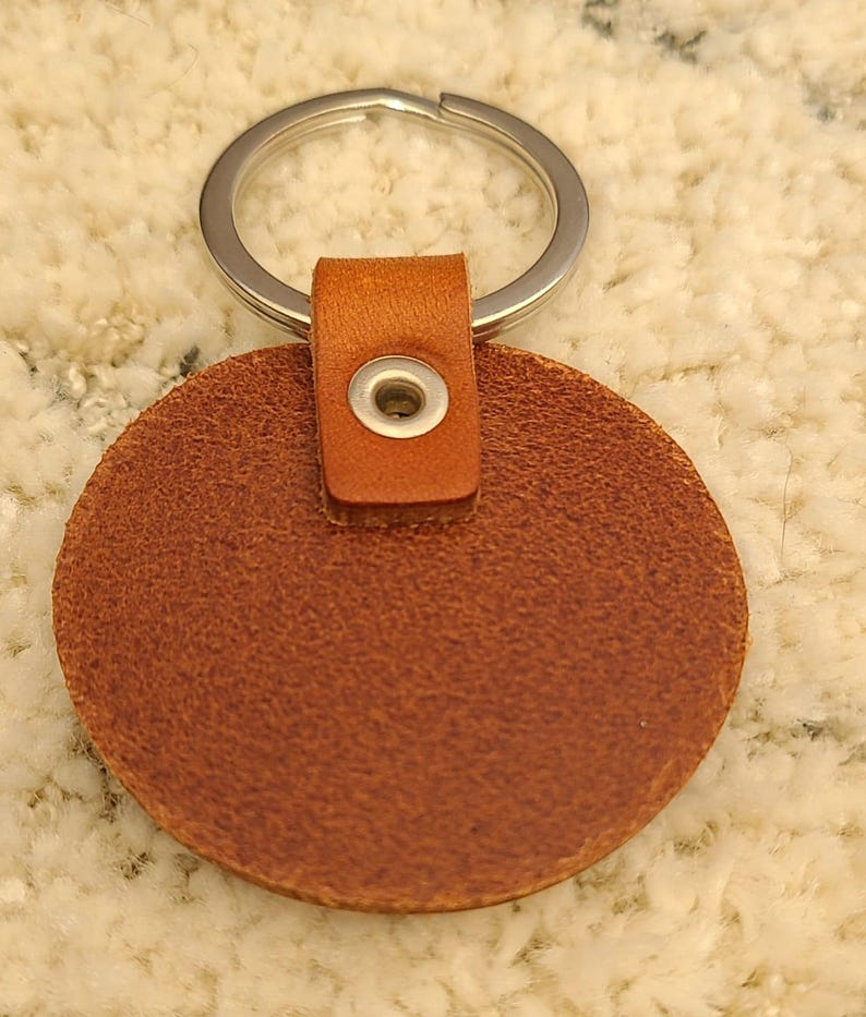 May include: A circular brown leather keychain featuring a textured surface and a silver metal split ring. The leather disc is attached to the ring by a folded leather tab secured with a small, centered silver metal eyelet grommet.