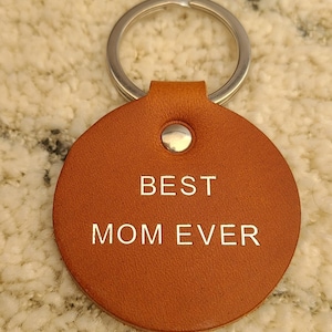 May include: A round, tan leather keychain featuring a silver metal ring at the top. The leather disc is secured with a silver rivet and has the words BEST MOM EVER embossed in a clean, sans-serif font across the center of the circular surface.