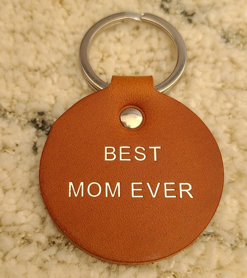 May include: A round, tan leather keychain featuring a silver metal ring at the top. The leather disc is secured with a silver rivet and has the words BEST MOM EVER embossed in a clean, sans-serif font across the center of the circular surface.