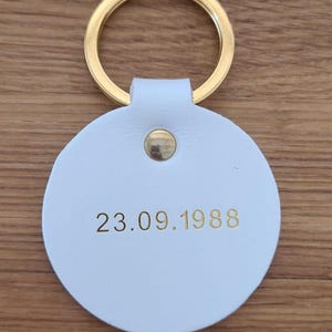 May include: A circular white keychain featuring a gold-colored metal ring and a central gold rivet. The date 23.09.1988 is embossed in gold lettering on the front surface of the white circular tag, which is attached to the metal ring.