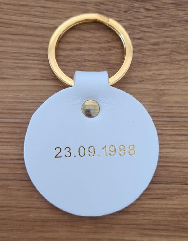 May include: A circular white keychain featuring a gold-colored metal ring and a central gold rivet. The date 23.09.1988 is embossed in gold lettering on the front surface of the white circular tag, which is attached to the metal ring.