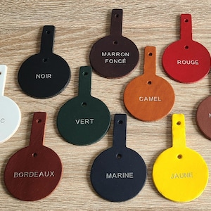 May include: Ten circular leather tags with tabs, each featuring a single hole and a stamped color name. The colors include white, black, dark brown, red, green, camel, light brown, burgundy, navy blue, and yellow, arranged on a flat surface.