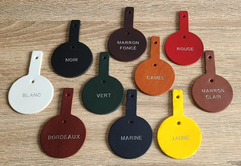 May include: Ten circular leather tags with tabs, each featuring a single hole and a stamped color name. The colors include white, black, dark brown, red, green, camel, light brown, burgundy, navy blue, and yellow, arranged on a flat surface.