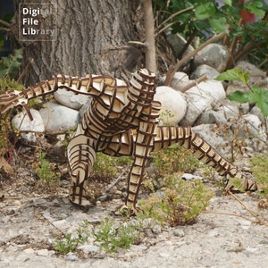 May include: A wooden sculpture of a person in a crouching pose. The sculpture is made of many interlocking pieces of wood and is painted brown. The sculpture is standing on a bed of gravel and green plants.