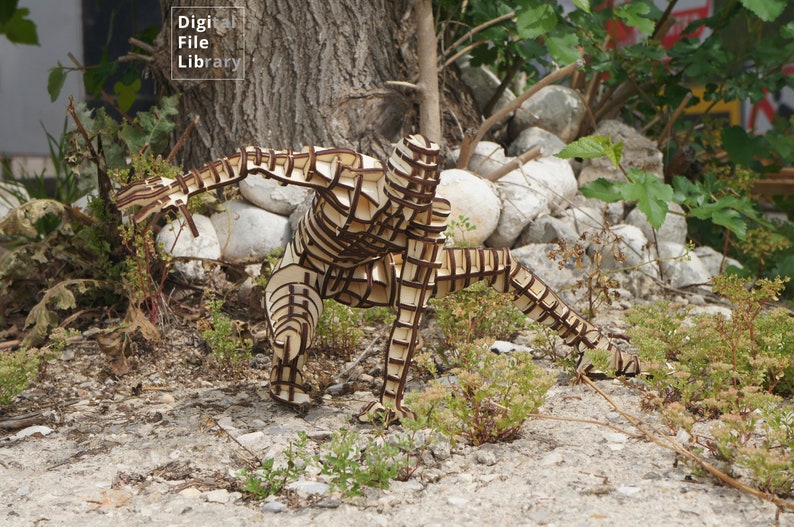 May include: A wooden sculpture of a person in a crouching pose. The sculpture is made of many interlocking pieces of wood and is painted brown. The sculpture is standing on a bed of gravel and green plants.