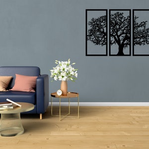 K&ouml;nnte beinhalten: Ein blaues Sofa mit Kissen steht vor einer grauen Wand mit einem schwarzen Wandbild aus Metall, das eine Baumsilhouette zeigt. Ein kleiner goldener Beistelltisch mit einer Vase mit wei&szlig;en Blumen und einer Uhr steht neben dem Sofa. Ein goldener Couchtisch aus Metall steht vor dem Sofa.
