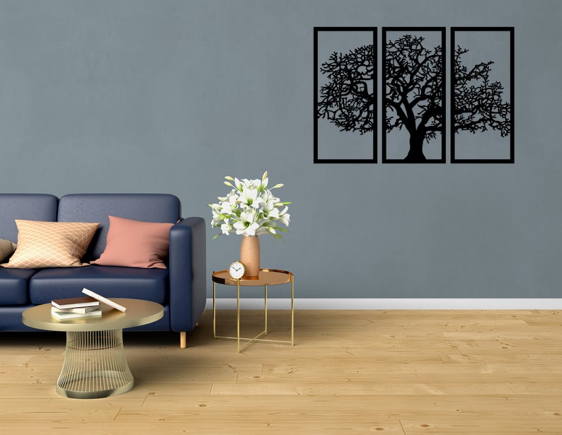 K&ouml;nnte beinhalten: Ein blaues Sofa mit Kissen steht vor einer grauen Wand mit einem schwarzen Wandbild aus Metall, das eine Baumsilhouette zeigt. Ein kleiner goldener Beistelltisch mit einer Vase mit wei&szlig;en Blumen und einer Uhr steht neben dem Sofa. Ein goldener Couchtisch aus Metall steht vor dem Sofa.