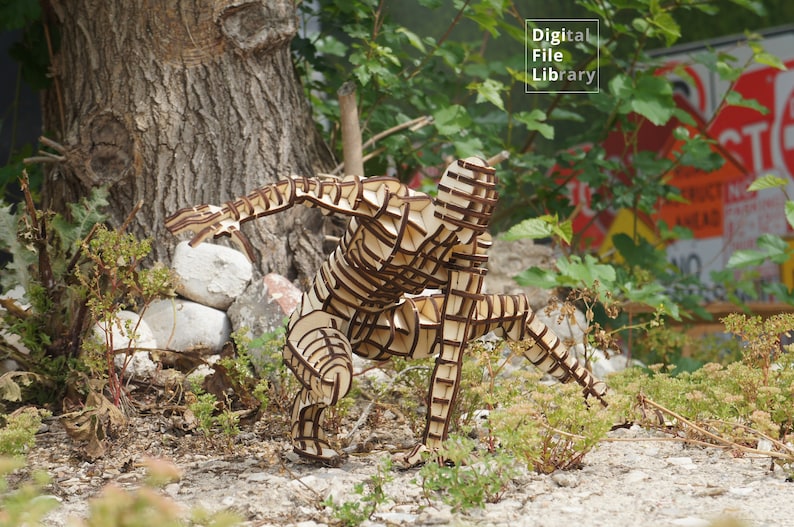 May include: A wooden sculpture of a person in a crouching pose. The sculpture is made of many interlocking pieces of wood, creating a lattice-like effect. The sculpture is standing on a bed of brown gravel.