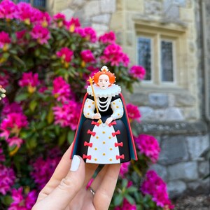 K&ouml;nnte beinhalten: Eine kleine Figur einer K&ouml;nigin, die ein schwarz-wei&szlig;es Kleid mit roten Schleifen und einer goldenen Krone tr&auml;gt. Sie h&auml;lt ein Zepter in ihrer rechten Hand. Die Figur wird vor einem Steinbau mit rosa Blumen gehalten.
