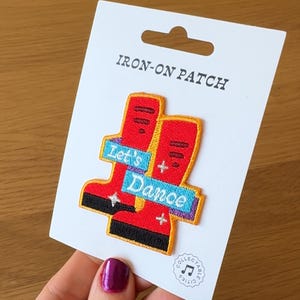 Könnte beinhalten: Ein Bügelbild mit roten Stiefeln und dem Schriftzug "Let's Dance" auf einem blauen Banner. Das Bügelbild befindet sich auf einer weißen Karte mit dem Text "IRON-ON PATCH" oben.
