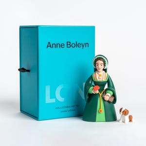 Könnte beinhalten: Eine türkisfarbene rechteckige Schachtel mit der Aufschrift Anne Boleyn und LON Collectable Art Toy second edition, dazu eine Figur in einem dunkelgrünen Kleid, die eine rote Blume und ein Buch hält, sowie ein kleiner braun-weißer Hund.