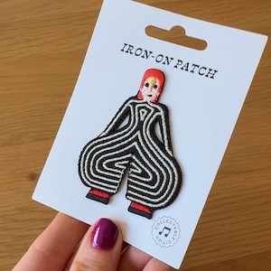 Puede incluir: Un parche termoadhesivo con una figura estilizada a rayas blancas y negras, con la cabeza y los pies rojos. El parche está sobre una tarjeta blanca que dice "IRON-ON PATCH". La parte inferior de la tarjeta tiene un logotipo circular con notas musicales.