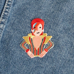 May include: An embroidered patch featuring a stylized figure with bright red hair, a blue lightning bolt across the face, and a gold, black, and red striped outfit with gold shoulder accents. The patch is attached to a blue denim fabric background.