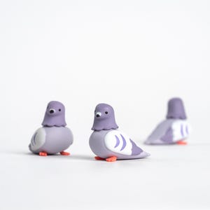 May include: Three small, stylized pigeon figurines. The birds have lavender bodies, grey heads, and orange feet. Each has a white patch with curved lines on its wings. The figurines are set against a white background.