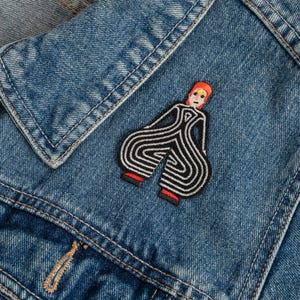 Puede incluir: Una chaqueta de mezclilla azul con un parche bordado de una figura estilizada con cabello naranja, rostro pálido y un atuendo de rayas blancas y negras con piernas anchas y curvas. La figura tiene pequeños detalles rojos en los pies y un círculo.